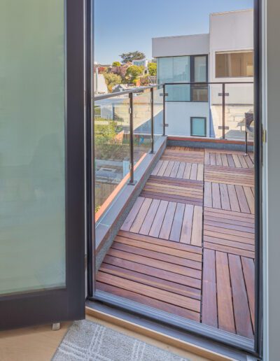 View from an open door leading to a modern wooden deck balcony with glass balustrade overlooking a residential area.