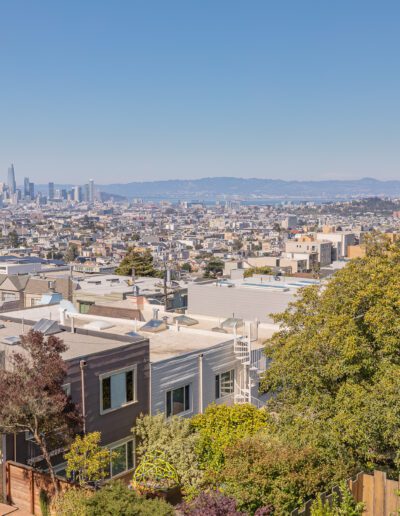 View of a cityscape with buildings and distant skyscrapers under a clear blue sky, as seen from an elevated residential area with trees and houses in the foreground.