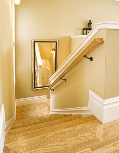 A well-lit staircase with wooden flooring, beige walls, and a handrail leading to a hallway with a mirror.