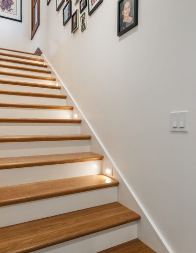 A wooden staircase with illuminated step edges, white walls adorned with framed pictures, leading upwards.